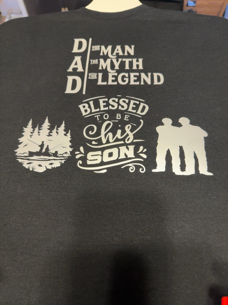Finished black Dad, the man, the myth, the legend shirt with Blessed to be his son lettering and cream father-and-son silhouettes, made by Chevelle’s Customs.